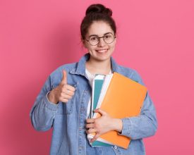 young-student-woman-wearing-denim-jacket-and-eyeglasses-holding-colorful-folders-and-showing-thumb-up-on-pink.jpg